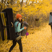 a woman wearing a backpack on her front and a crash pad on her back walking to a bouldering area in the fall