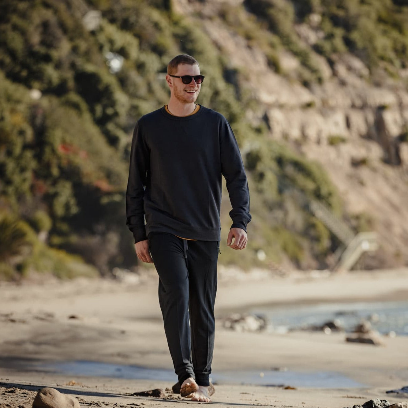 A man smiles wearing sunglasses and the Ridge Natural Tencel Crew Sweatshirt walking on a beach